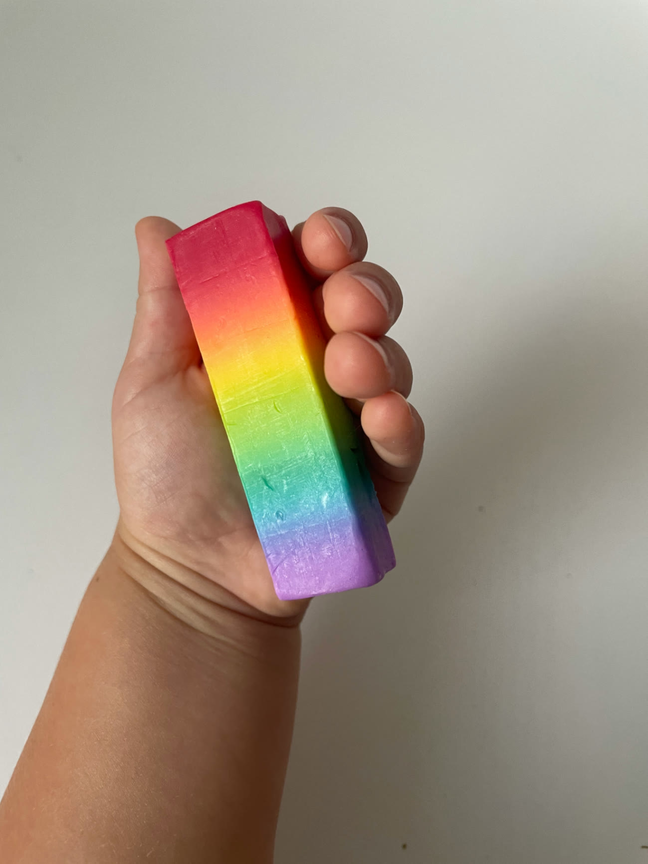 A child's hand holding a tall rainbow-layered Kind & Fern soap bar